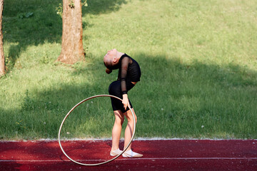 A girl in a black outfit performs a backbend pose with a hoop on an outdoor track. The green grass and natural surroundings enhance the focus on her graceful gymnastics practice.