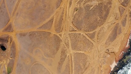 A vertical drone reveals a coastal desert webbed with tire tracks as teal surf slams black rocks, blending human tracery with raw shoreline textures and wind-hardened stillness. - Powered by Adobe