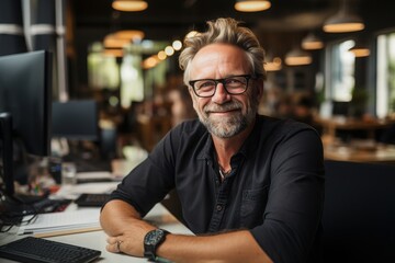 A man with glasses and a beard sits at a desk in a contemporary office, exuding confidence and warmth. Natural light brightens the open workspace around him