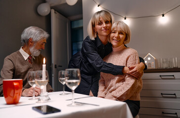 A family shares a warm moment during a festive dinner. A woman embraces another, while an elder sits nearby. The cozy setup features string lights, a lit candle, and elegant tableware.