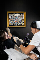 Tattoo artist working on a client’s arm in a studio, with framed tattoo designs on the wall. Concept of body art, lifestyle, creativity, and modern tattoo culture.