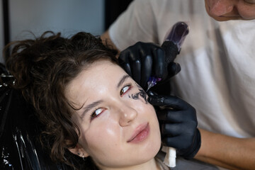 Smiling young woman getting a face tattoo in a modern studio, expressing confidence and joy during...