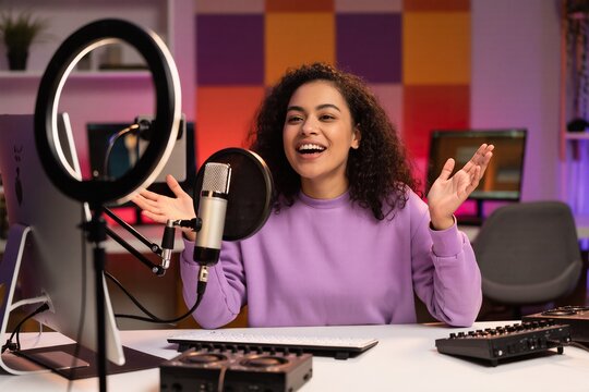 A cheerful woman with diverse ethnic features smiles brightly while speaking into a sleek microphone during a live podcast session, surrounded by modern studio equipment