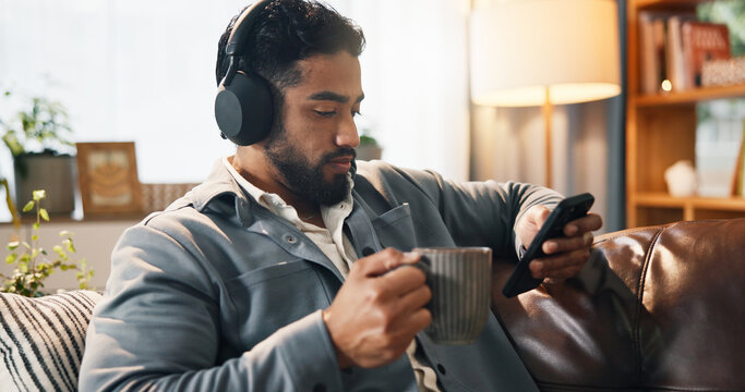 Coffee, man and phone with headphones in home for music playlist, social media video or podcast. Online, person and relax with smartphone on sofa for listening to radio, album and audio subscription - Powered by Adobe