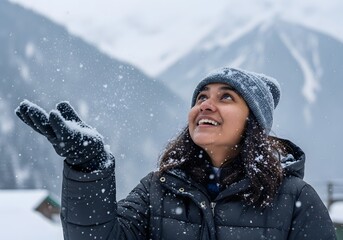 A joyful young woman in a winter coat and hat smiles as she looks up, catching snowflakes in her hand during a snowfall in a beautiful mountain landscape