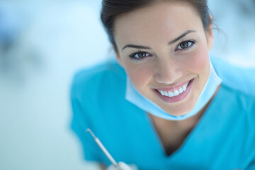 Smiling young female dental professional in blue scrubs with mask under chin holding dental tool