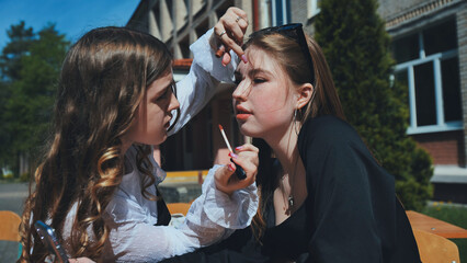 Two schoolgirls applying makeup during a sunny school day, sitting together on a bench outdoors, enjoying their friendship and fun