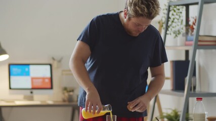 Young Caucasian man standing in at table pouring orange juice in glass, drinking it and watching something on smartphone - Powered by Adobe