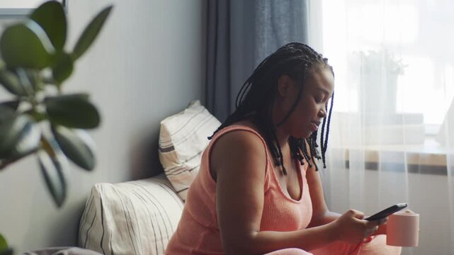 Horizontal side view shot of modern young Caucasian woman sitting relaxed on bed in bedroom surfing social net on smartphone