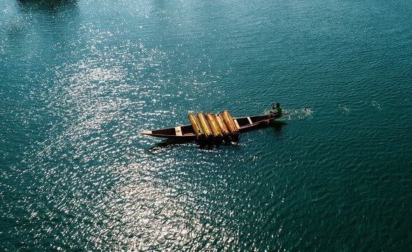 Aerial view of a traditional wooden boat gliding across the shimmering, sun-drenched waters, carrying stacks of bamboo, creating a striking contrast against the deep blue, Location Data Omitted.