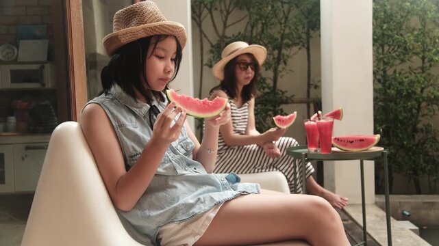 Chilling Asian family holidays, casual girl and mum enjoy eating pieces of fresh red watermelons sliced and smoothie drinks, happiness relaxing on vacation, and hot summertime at outdoor terrace.