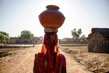 Woman Balancing Pot on Head Walks Through Rural Village Landscape