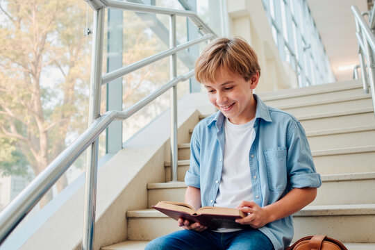Caucasian boy sitting on staircase reading book and smiling, light brown hair and casual clothing, natural light streaming through large windows in modern building interior - Powered by Adobe