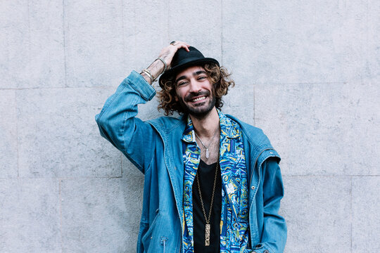 Smiling young man touching hat while standing in front of wall