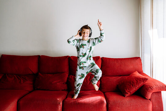 Happy boy listening to music through headphones while dancing on sofa against wall