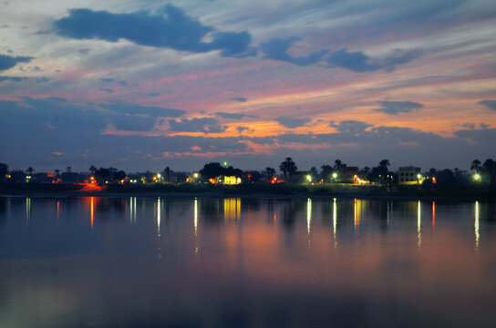 View of the tranquil river reflecting the soft, glowing lights of the distant shore under a sky brushed with hues of twilight, Philae Temple, Luxor Governorate, Egypt.