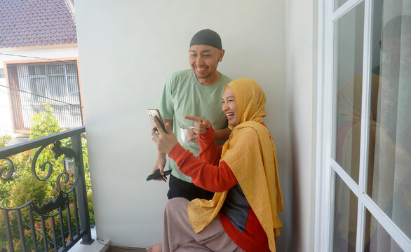Happy Asian Muslim Couple Having Video Call On Smartphone While Enjoying Coffee At Balcony