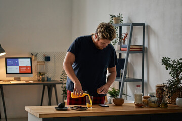 Horizontal medium shot of young Caucasian man standing at table at home pouring juice into glass and watching something on smartphone