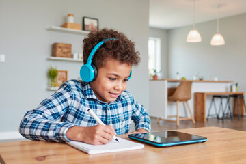 Black boy child wearing headphones sitting at table smiling while writing in notebook and looking at tablet screen, engaging in remote learning or online education at home