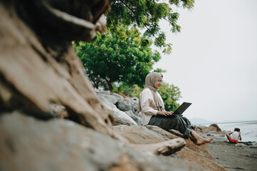 Muslim mother working on laptop on the beach with children playing near the shore
