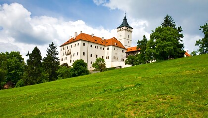 Fototapeta premium Majestic white castle with orange roof and spire, set against a green grassy hill and cloudy sky.