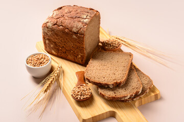 Wooden board with sliced loaf of bread, wheat spikelets and grains on grey background