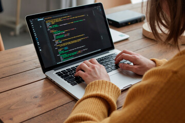 Caucasian young adult woman typing on laptop computer displaying programming code, sitting at wooden table, focusing on software development task, hands on keyboard, partial face visible