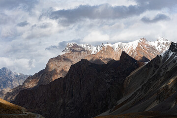 Obraz premium Dramatic sunlight breaks through clouds, casting a golden glow on a majestic mountain range while others remain in deep shadow. This high-contrast, epic landscape evokes power and awe.