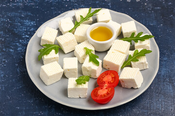 Plate with tasty cut feta cheese, tomatoes and arugula on blue grunge background