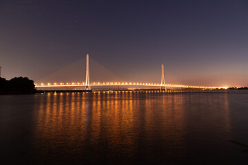 Fototapeta premium Luminous suspension bridge stretching across a dark expanse of water under a twilight sky. The bridge's lights create a stunning reflection on the calm surface below.