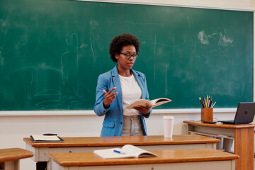 Black woman standing in classroom holding open book and gesturing while teaching in front of chalkboard with laptop and coffee cup on desk in background