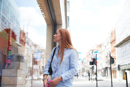 Young redhead woman looking through store window in city