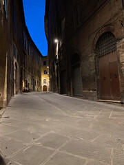 Fototapeta premium Narrow cobblestone street in Siena, Italy, illuminated by streetlights at dusk, creating a captivating blue hour atmosphere