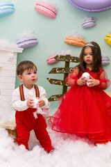 A little girl and a boy in red clothes have fun in the artificial snow by the fireplace in the studio. The girl is holding a box with gifts. Little kids in red christmas costume having fun beside a