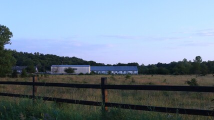 A long white farm building in the background stands on a dry grassy field behind a low wooden fence, a farm landscape with cowsheds, barns, and meadows for grazing farm animals.