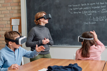 Caucasian middle aged woman standing in front of chalkboard instructing two Caucasian children wearing virtual reality headsets, boy gesturing with hand while girl sitting at table
