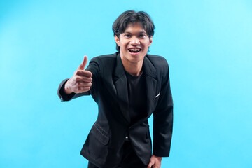 A confident young Asian man in a black suit smiles brightly while showing a thumbs-up gesture, expressing approval, enthusiasm, and positive energy in a professional manner.