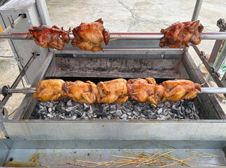 Juicy whole chicken are roasting on a rotisserie over charcoal at a street vendor stall. The smell of grilled meat fills the air, attracting customer in a lively market atmosphere.