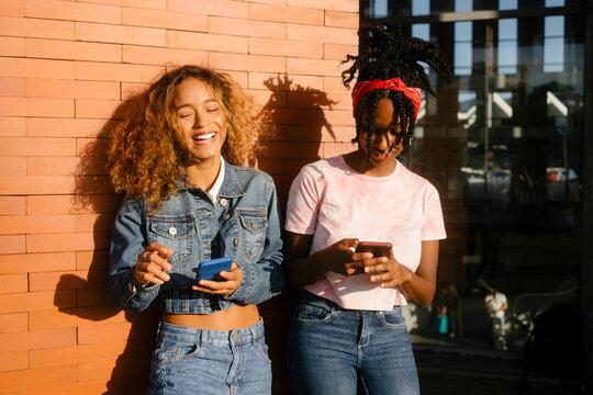 Young woman laughing by friend using mobile phone on sunny day