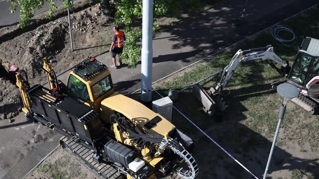 Construction worker in orange vest supervising excavator and drilling machine at city site with dug ground and equipment around showing teamwork and modern urban infrastructure development