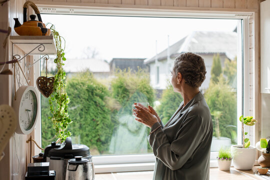 Mature woman holding tea glass at the window in kitchen