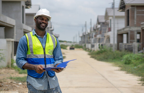 A construction worker in a safety vest and helmet is examining blueprint at a residential building site. Newly built structure surround him, showcasing ongoing construction work.