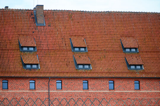 Historic building featuring red bricks and sloped tiled roof. Dormer windows break uniformity, offering contrast. Bright daylight enhances warm tones