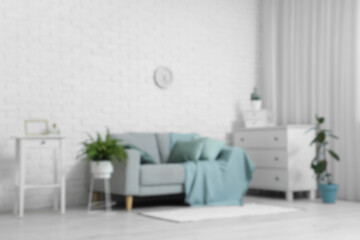 Grey sofa, houseplants, chest of drawers and clock on white brick wall in stylish living room, blurred view