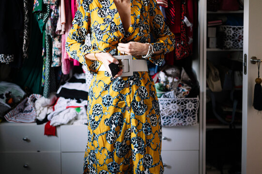 Mid adult woman fastening belt on new yellow dress in wardrobe at home