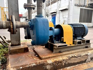 The image shows an industrial pump with a blue casing and a blue and yellow motor. The machinery is positioned on a rusty concrete base, connected to a metal pipe with a flange, highlighting an outdoo