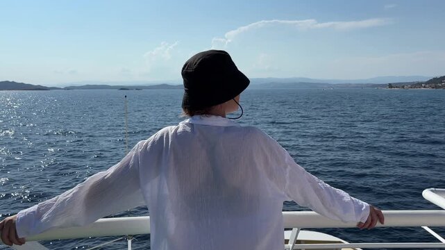 a young woman in a black bucket hat and white shirt is standing on a boat railing, looking out at the vast blue sea and distant hills, concept of travel, sailing, and tourism