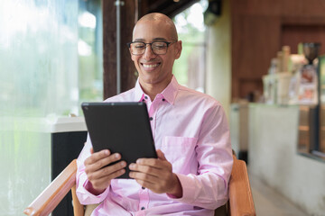 Bald mature Hispanic man wearing pink casual business shirt in coffee shop using tablet computer