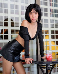 A stylish woman in a black off-shoulder top and leather skirt leans confidently against a table, holding a colorful drink in a modern outdoor setting with a grid-patterned background