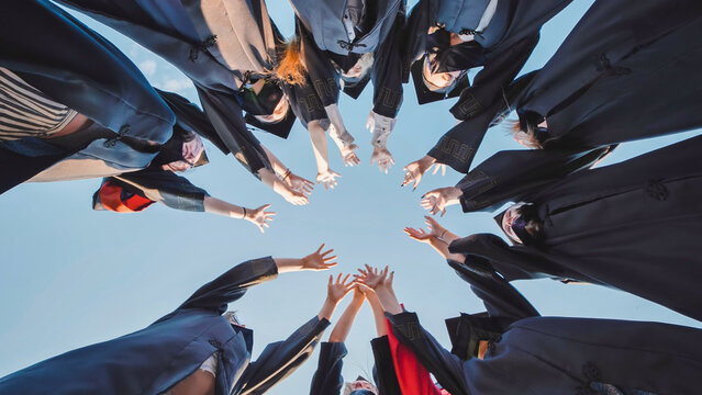 Group of students in graduation attire holding hands in a circle against a clear blue sky, commemorating their graduation